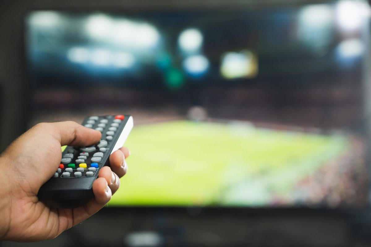 Person richtet die Fernbedienung auf einen Fernseher. Auf dem Bildschirm wird ein Fußballfeld angezeigt.