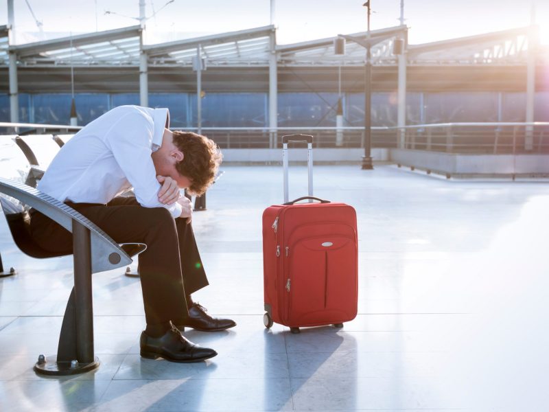 Mann sitzt in verzweifelter Haltung auf einem Flughafen. Neben ihm steht ein Koffer.