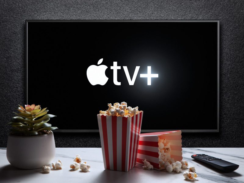 Apple TV+ wird auf einem Fernseher angezeigt. Auf einem Tisch davor steht Popcorn.