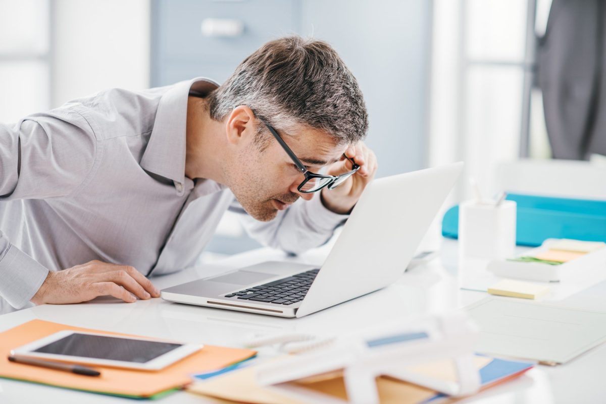 Mann mit Brille guckt intensiv auf einen Laptop, der auf einem Schreibtisch mit mehreren Dokumenten liegt.