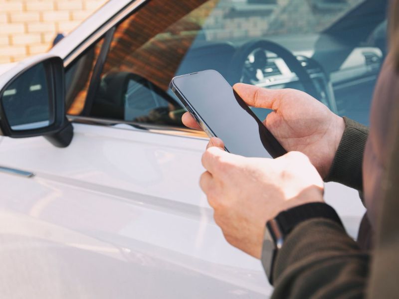 Person steht mit Smartphone in der Hand vor einem Auto.