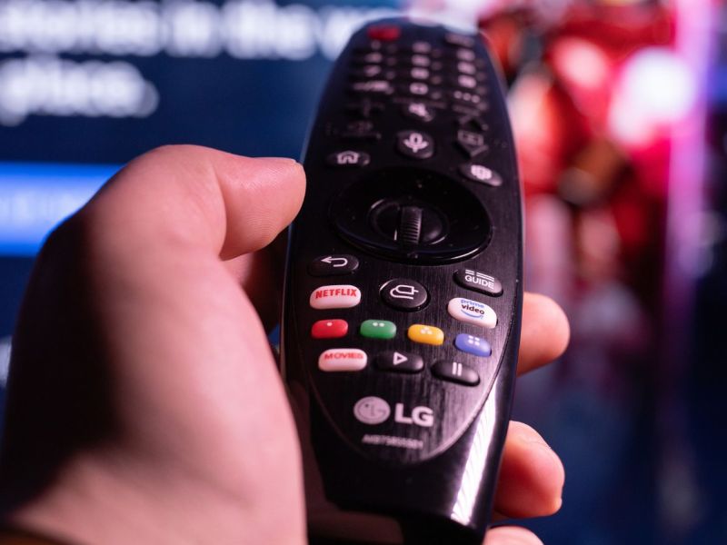 Person hält eine Fernbedienung in der Hand, die in Richtung eines Fernsehers zeigt.