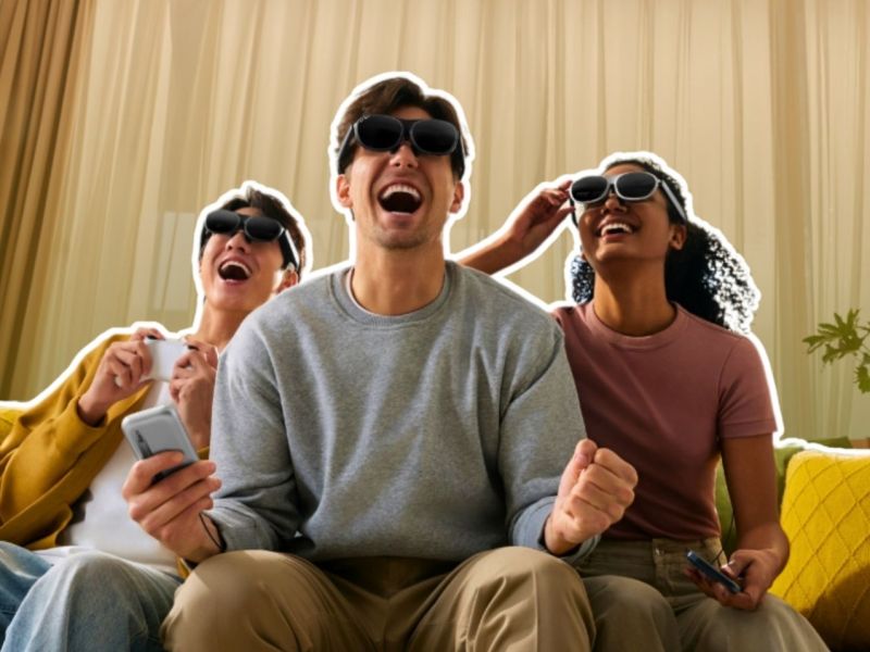 Drei junge Erwachsene sitzen lachend auf einem Sofa und tragen Rokid-AR-Brillen. Sie halten Smartphones / Controller in den Händen und jubeln, als würden sie gemeinsam ein Spiel spielen oder einen Film schauen.