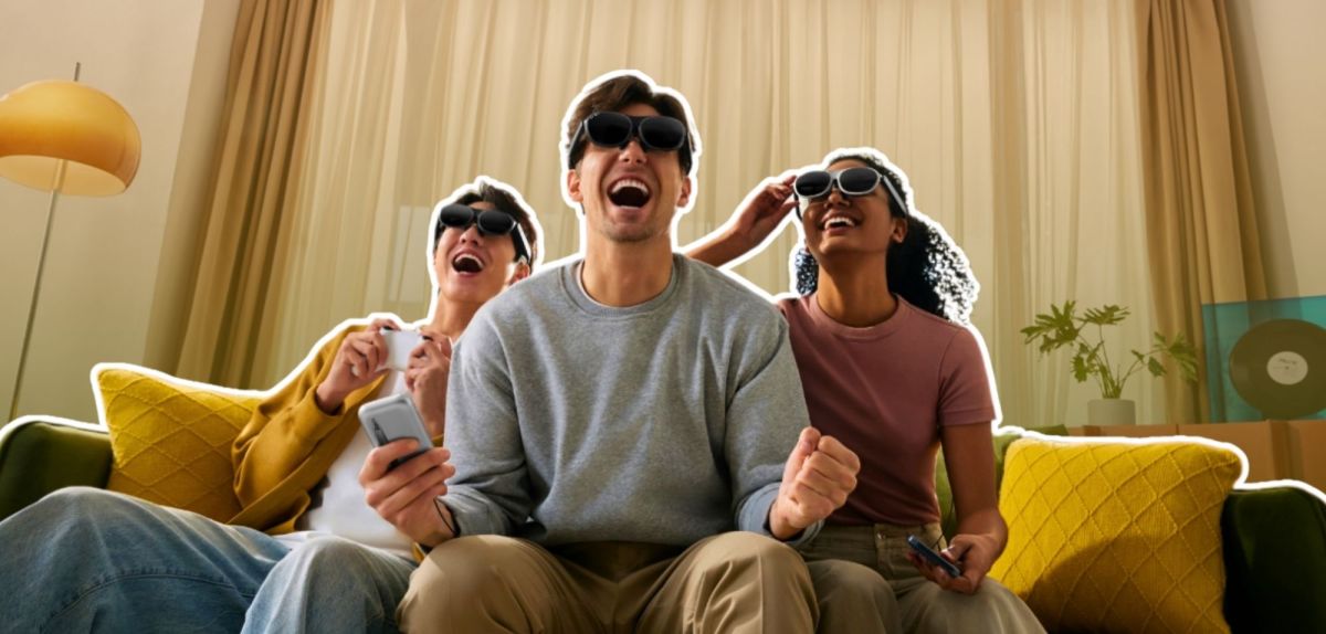 Drei junge Erwachsene sitzen lachend auf einem Sofa und tragen Rokid-AR-Brillen. Sie halten Smartphones / Controller in den Händen und jubeln, als würden sie gemeinsam ein Spiel spielen oder einen Film schauen.